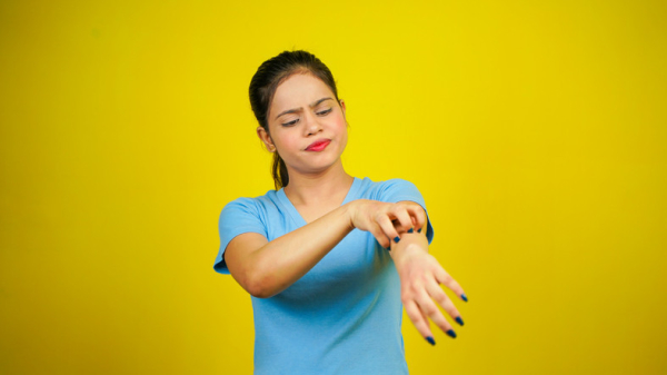 Woman with dark hair and a blue shirt itching her extended arm; background is yellow